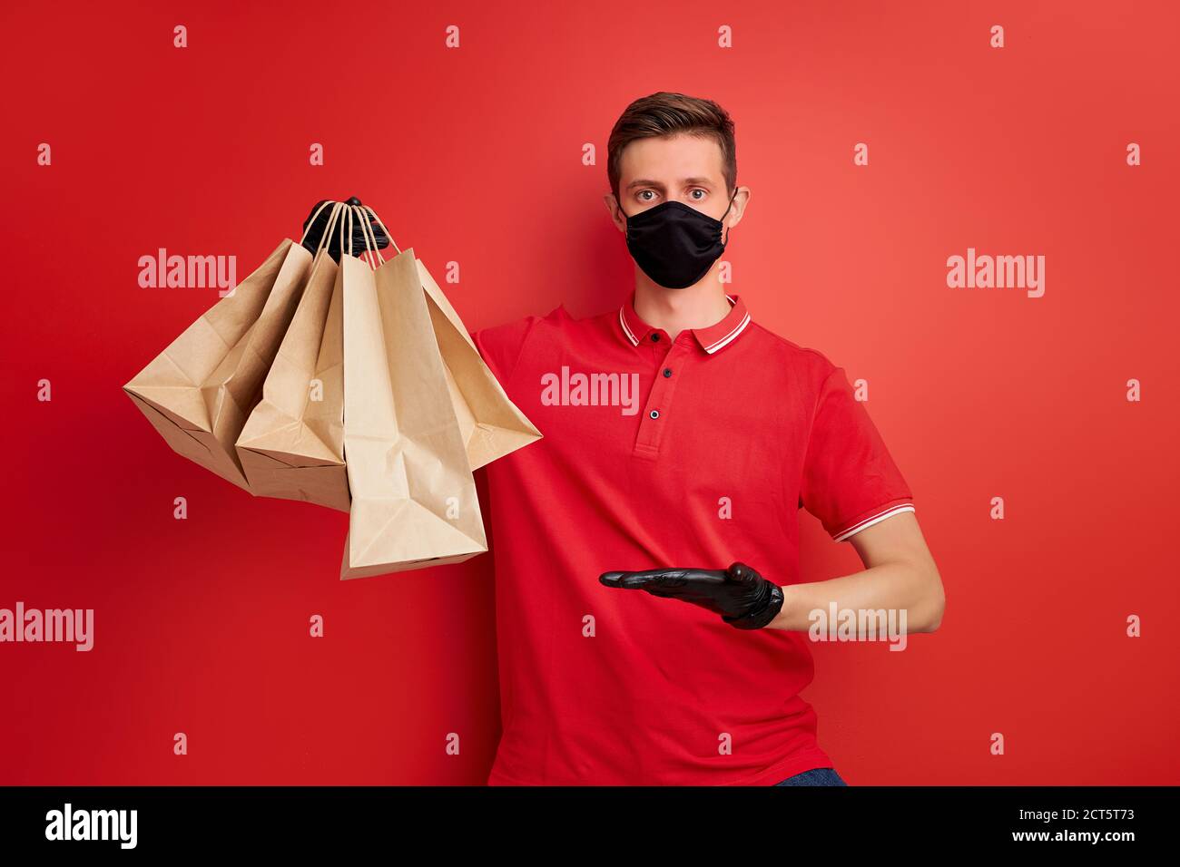 jeune homme de livraison caucasien employé en t-shirt rouge uniforme et masque tenir le paquet de papier d'artisanat avec les aliments isolés sur fond rouge, quaranti de service Banque D'Images