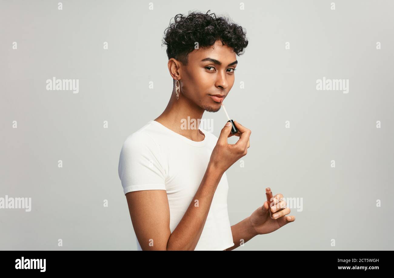 Jeune homme appliquant du brillant à lèvres. Homme liquide de genre mettant sur le maquillage sur ses lèvres avec applicateur contre fond blanc. Banque D'Images