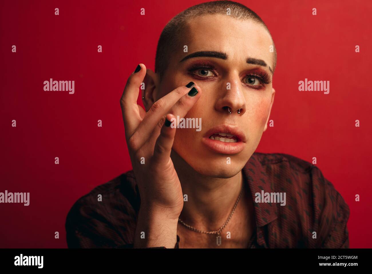 Homme transgenre mettant le maquillage sur fond rouge. Homme avec maquillage et perçage du nez regardant la caméra. Banque D'Images