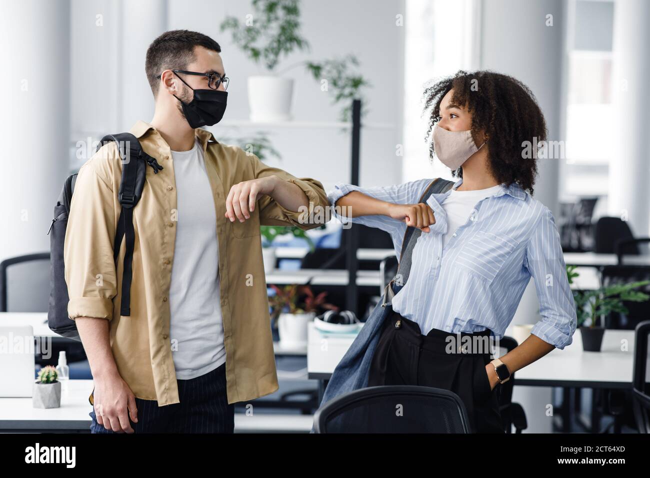 Nouvel accueil pour éviter la propagation du coronavirus. Guy et afro-américaine femme dans les masques protecteurs bonjour avec des coudes le matin à l'intérieur du bureau Banque D'Images