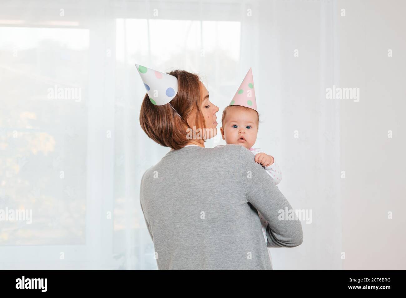 Une jeune mère tient un bébé mignon dans ses bras. Chapeaux en papier festif sur la tête. Vue arrière. Sur l'arrière-plan. Concept de maternité et heureux Banque D'Images