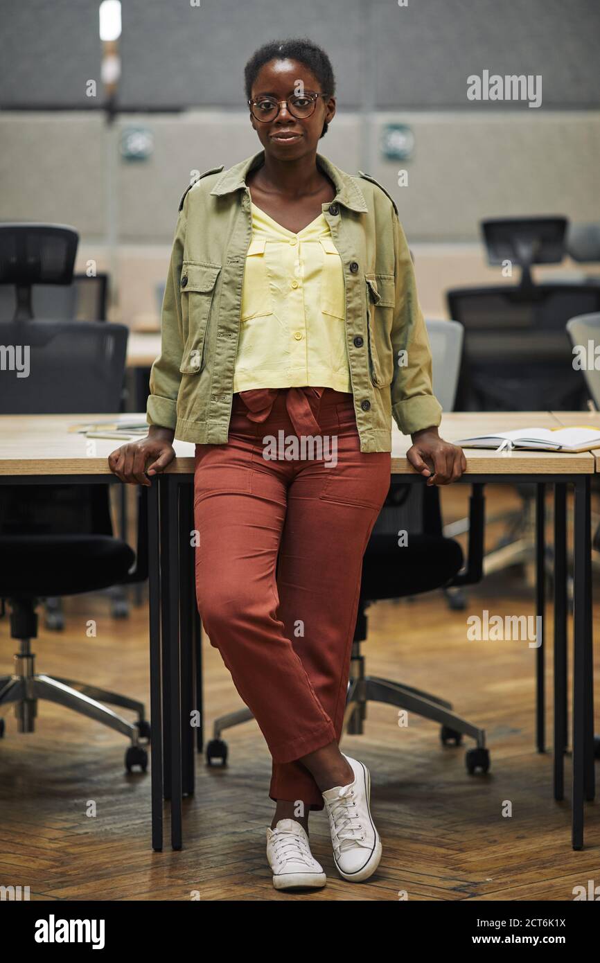 Portrait vertical de la jeune femme afro-américaine appareil photo tout en se tenant debout sur un bureau dans un bureau moderne Banque D'Images