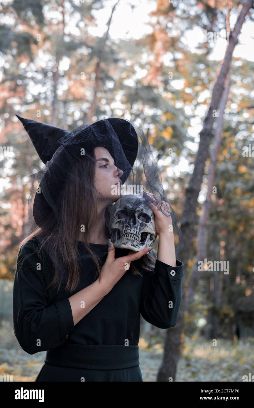 Jeune belle femme mignonne en robe sombre et chapeau de sorcière tient un crâne dans ses mains. Costume de fête d'Halloween. Forêt, parc avec arbres d'automne Banque D'Images