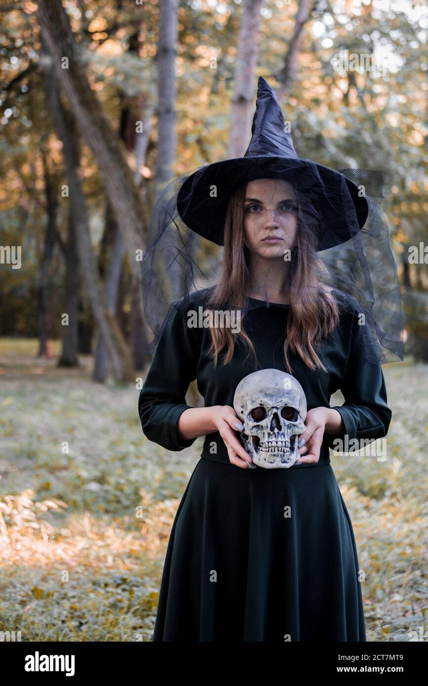 Jeune belle femme mignonne en robe sombre et chapeau de sorcière tient un crâne dans ses mains. Costume de fête d'Halloween. Forêt, parc avec arbres d'automne Banque D'Images