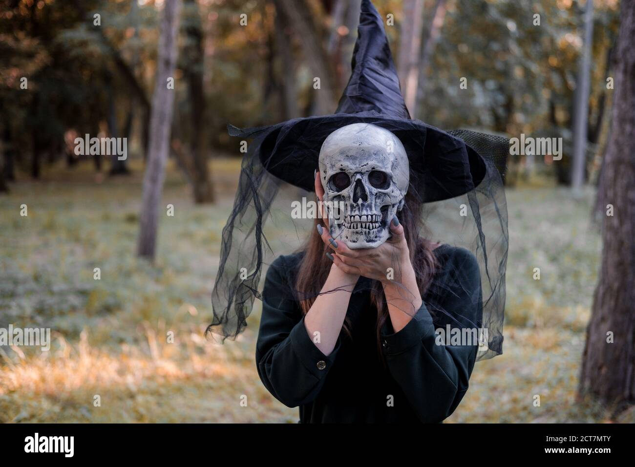Jeune belle femme mignonne en robe sombre et chapeau de sorcière tient un crâne dans ses mains. Costume de fête d'Halloween. Forêt, parc avec arbres d'automne Banque D'Images