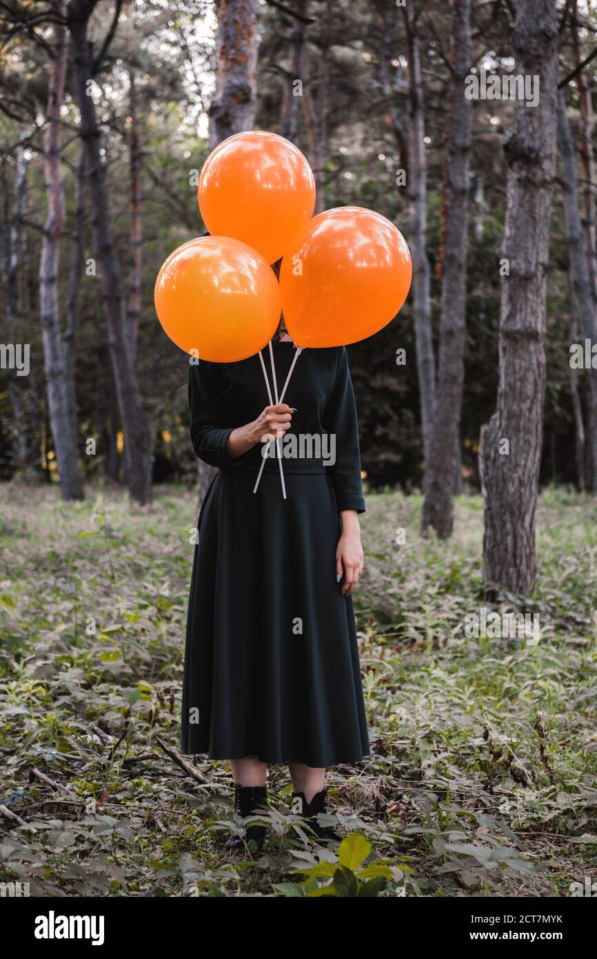 Jeune mignonne belle femme en robe sombre et chapeau de sorcière tient des ballons d'orange dans ses mains. Costume de fête d'Halloween. Forêt, parc avec arbres d'automne Banque D'Images