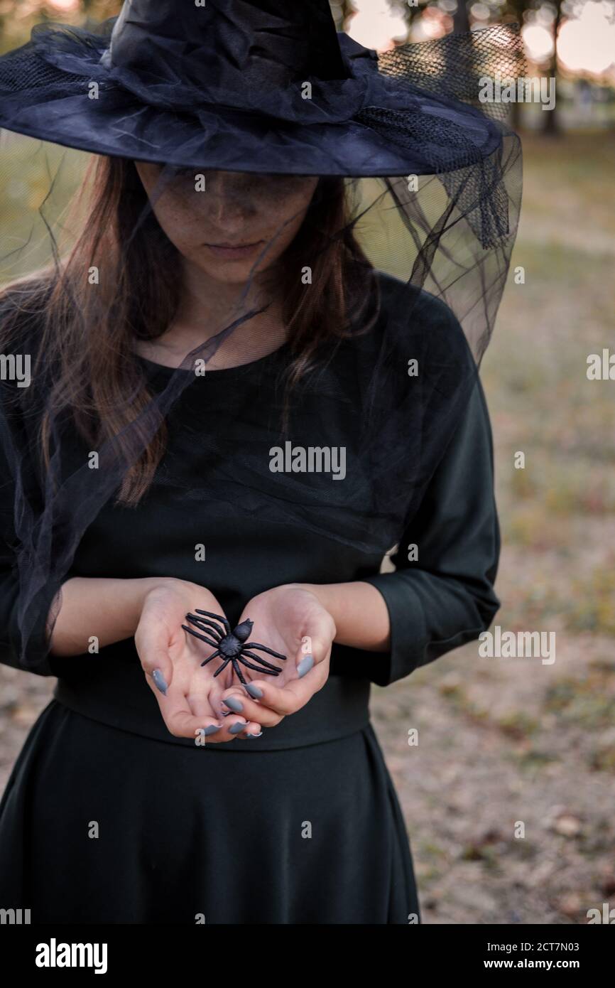 Jeune mignonne belle femme en robe sombre et chapeau de sorcière hotsblack araignée dans ses mains. Costume de fête d'Halloween. Forêt, parc avec arbres d'automne Banque D'Images