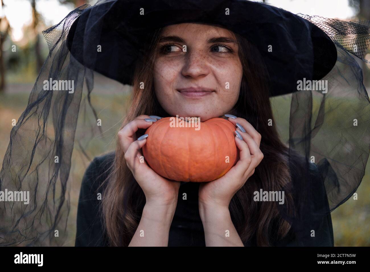 Une jeune femme en robe noire et chapeau de sorcière tient une citrouille dans ses mains. Costume de fête d'Halloween. Forêt, parc avec arbres d'automne Banque D'Images