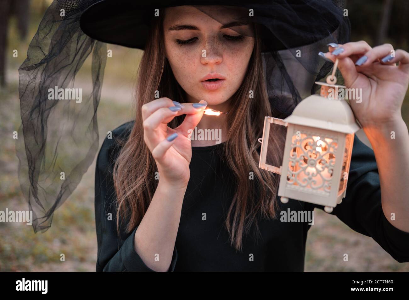 Une jeune femme en robe sombre et chapeau de sorcière tient une lanterne avec des bougies dans ses mains et illumine la forêt. Costume de fête d'Halloween. Garez-vous avec l'autom Banque D'Images