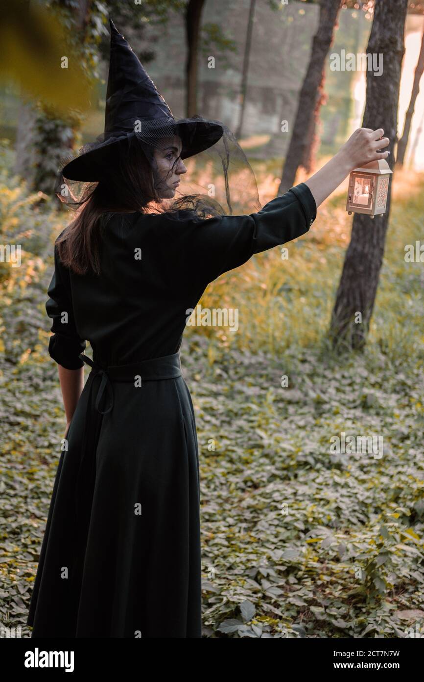 Une jeune femme en robe sombre et chapeau de sorcière tient une lanterne avec des bougies dans ses mains et illumine la forêt. Costume de fête d'Halloween. Garez-vous avec l'autom Banque D'Images