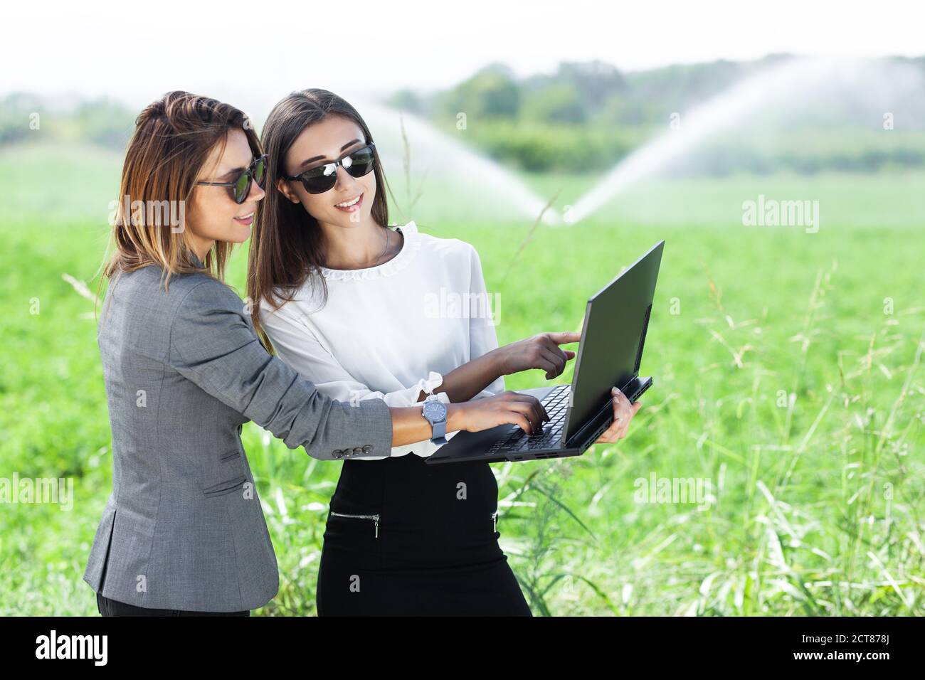 Femmes d'affaires avec un ordinateur portable dans un champ avec système d'irrigation agricole. Arroseurs d'eau en arrière-plan. Banque D'Images