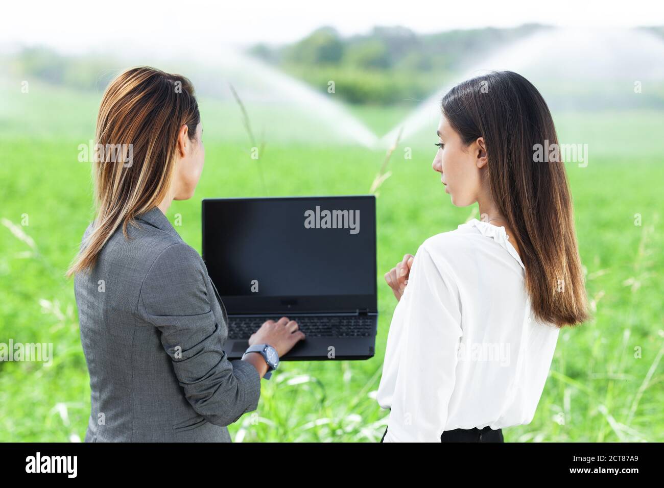 Femmes d'affaires avec un ordinateur portable dans un champ avec système d'irrigation agricole. Arroseurs d'eau en arrière-plan. Banque D'Images