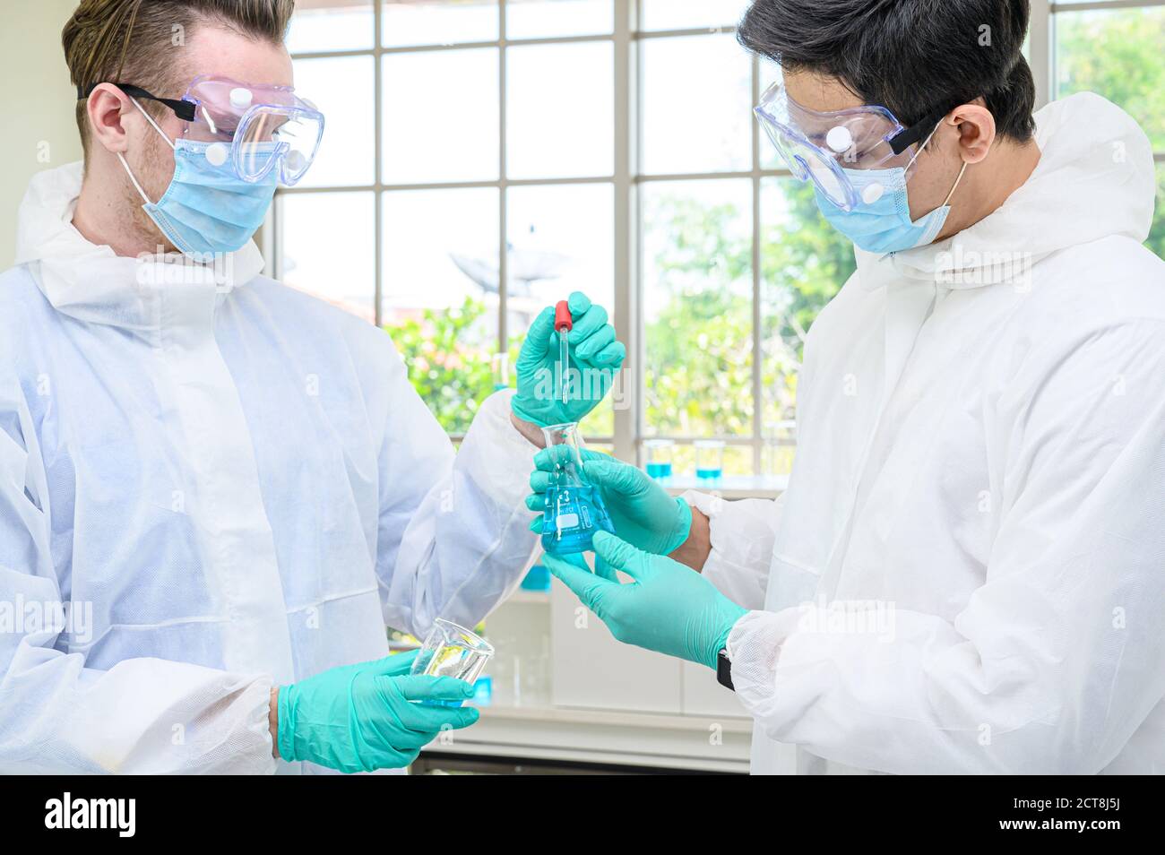 Couple scientifique mâle portant une combinaison de protection tenant le bécher ou le tube avec un échantillon de liquide à l'intérieur et en travaillant avec de nombreux équipements de laboratoire pour la recherche en laboratoire Banque D'Images