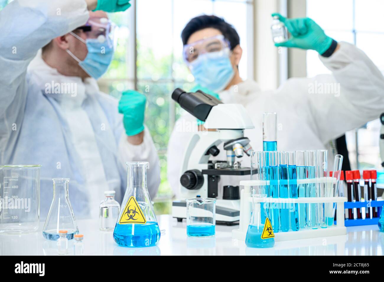 Équipement de laboratoire avec liquide bleu à l'intérieur du support sur la table avec couple scientifique mâle portant une combinaison de protection tenant le vaccin pour recherche en laboratoire Banque D'Images