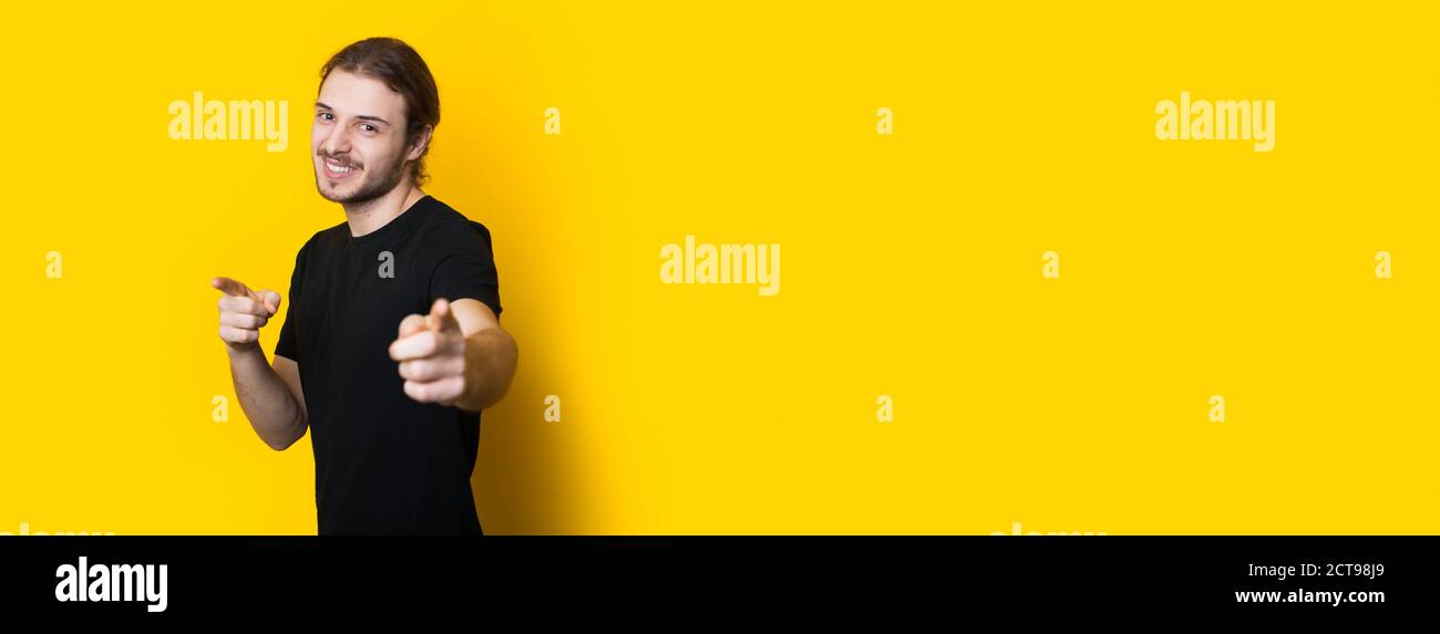 Homme caucasien barbu dans un t-shirt noir pointant vers l'appareil photo et souriez sur une bannière jaune studio Banque D'Images