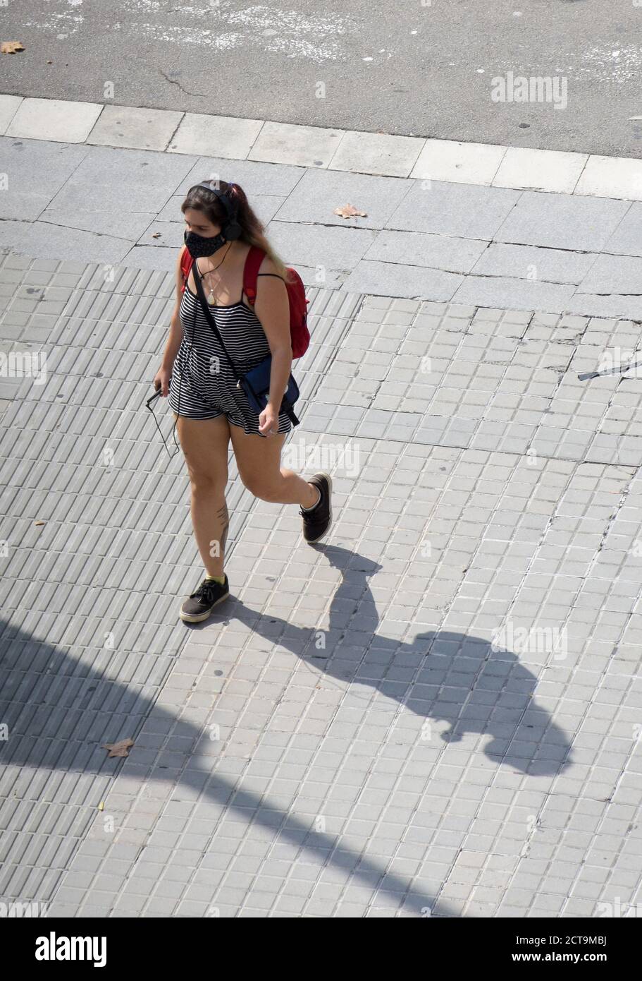 Une jeune femme avec un masque de protection marchant dans une rue pendant qu'elle écoute de la musique avec son mobile. Banque D'Images