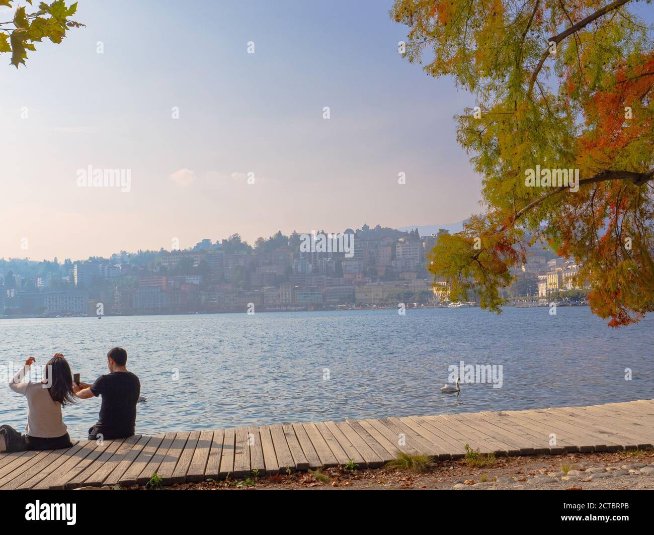 Jeune couple aimant assis sur la jetée en bois de Ciani Parc sur le lac de Lugano au coucher du soleil, vue sur le paysage urbain en automne Banque D'Images