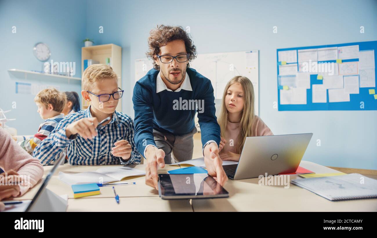 École primaire salle de classe: Un enseignant enthousiaste tenant un ordinateur tablette explique la leçon à des jeunes enfants brillants. Programmes d'apprentissage pour enfants Banque D'Images