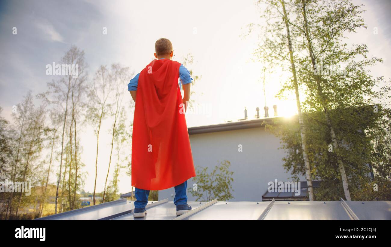 Garçon joue un rôle de Super Hero. Il est debout sur le toit d'une maison avec ses mains à la taille. Le jeune homme porte une cape rouge vif. Il l'est Banque D'Images