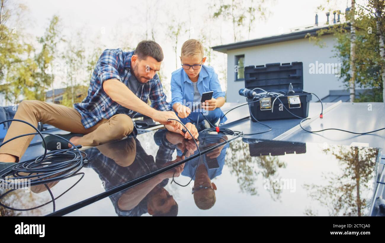 Père et fils installant des panneaux solaires sur une base métallique. Ils travaillent avec le câblage sur un toit de maison lors d'une journée ensoleillée. Garçon regarde un manuel sur un Banque D'Images