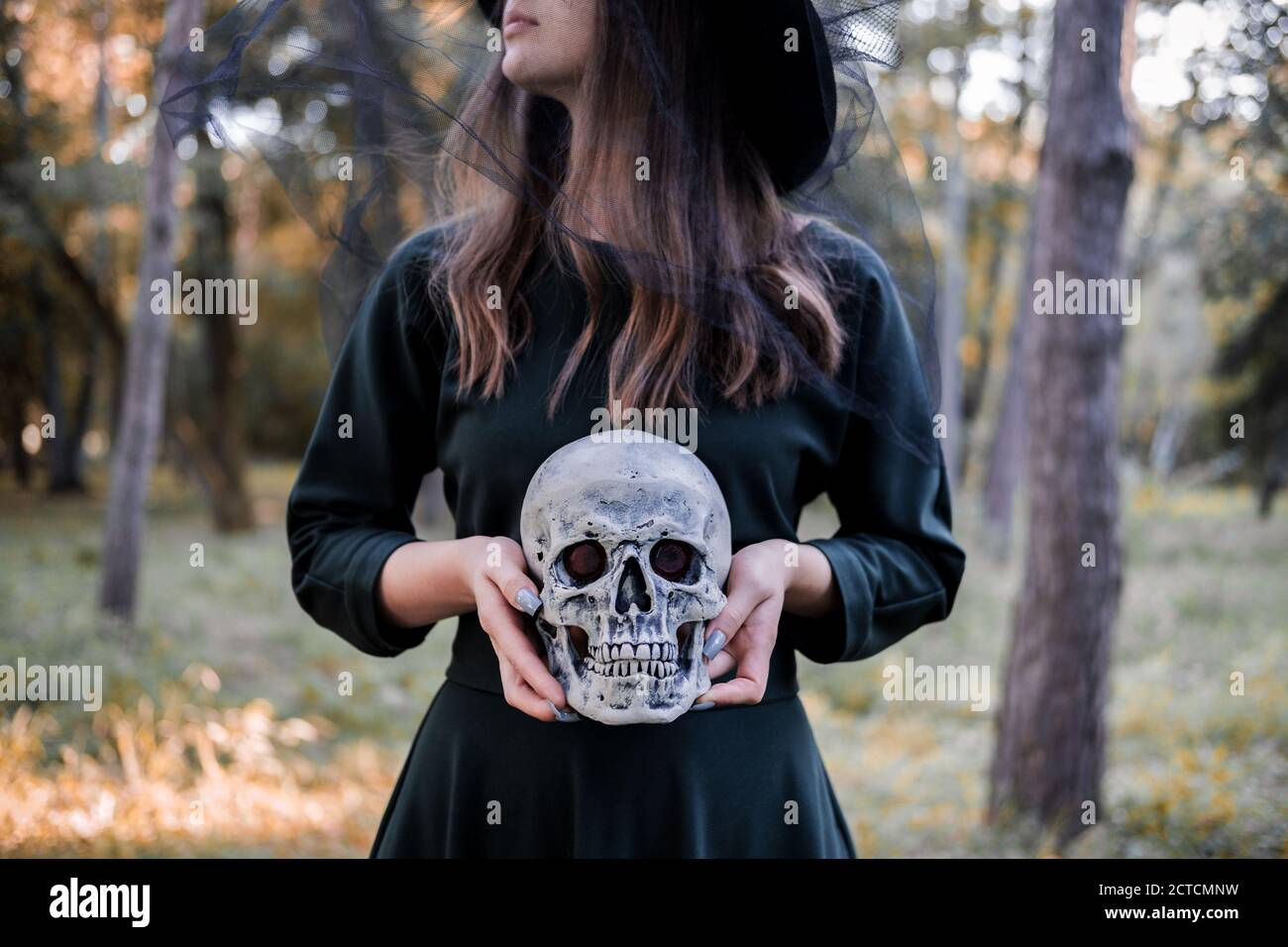 Jeune belle femme mignonne en robe sombre et chapeau de sorcière tient un crâne dans ses mains. Costume de fête d'Halloween. Forêt, parc avec arbres d'automne Banque D'Images