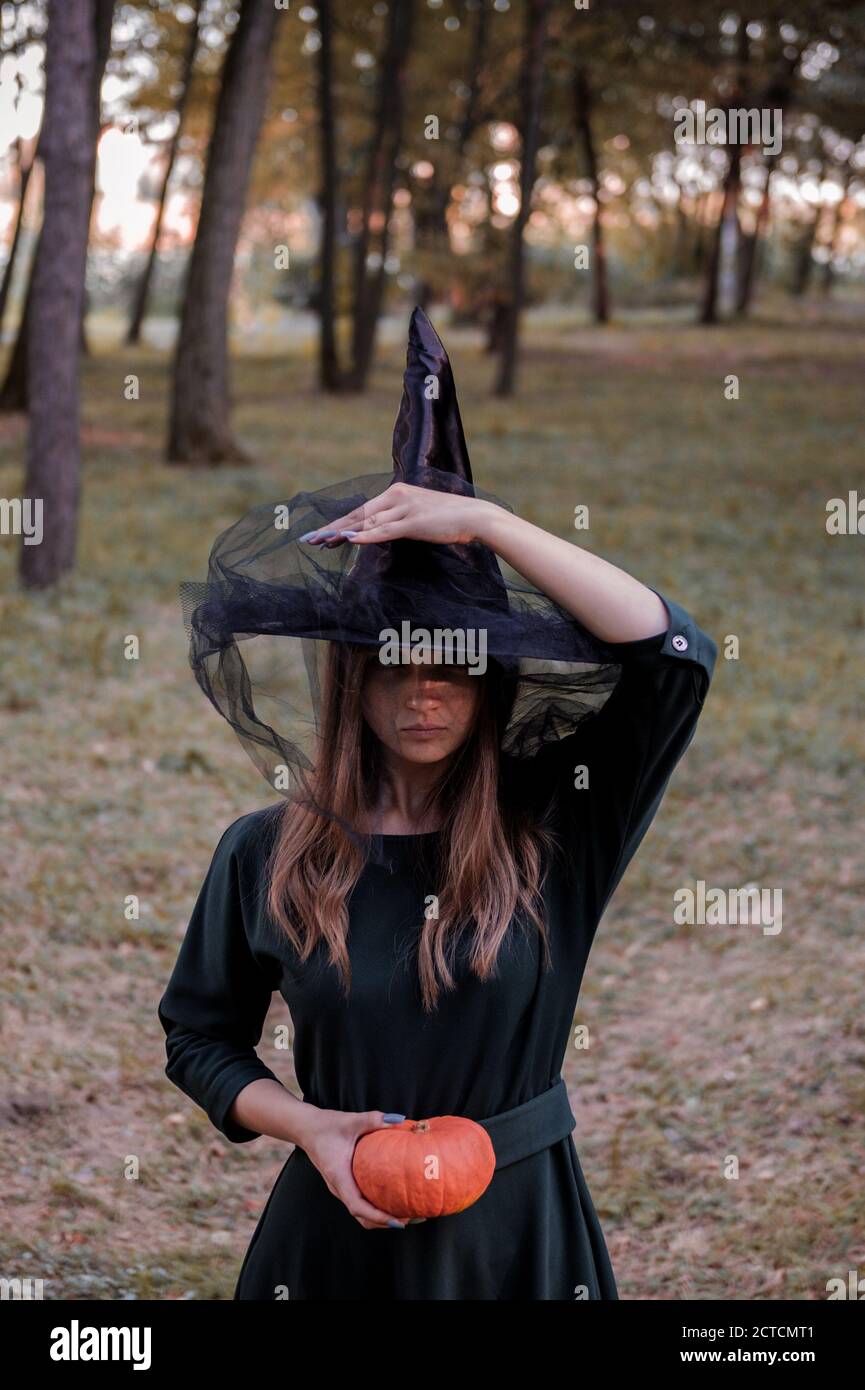 Jeune mignonne belle femme en robe sombre et chapeau de sorcière tient une citrouille orange dans ses mains. Costume de fête d'Halloween. Forêt, parc avec arbres d'automne Banque D'Images
