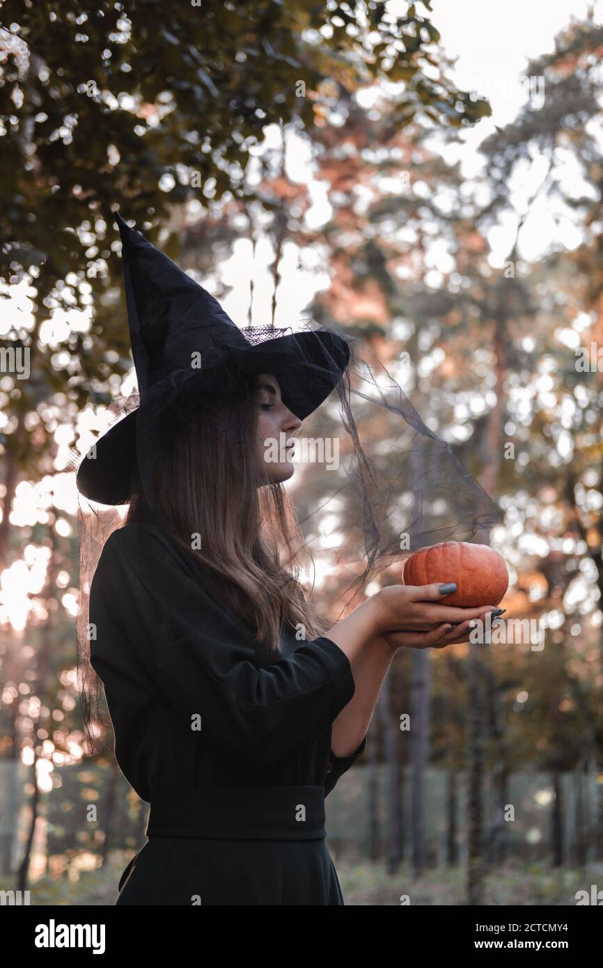 Jeune mignonne belle femme en robe sombre et chapeau de sorcière tient une citrouille orange dans ses mains. Costume de fête d'Halloween. Forêt, parc avec arbres d'automne Banque D'Images
