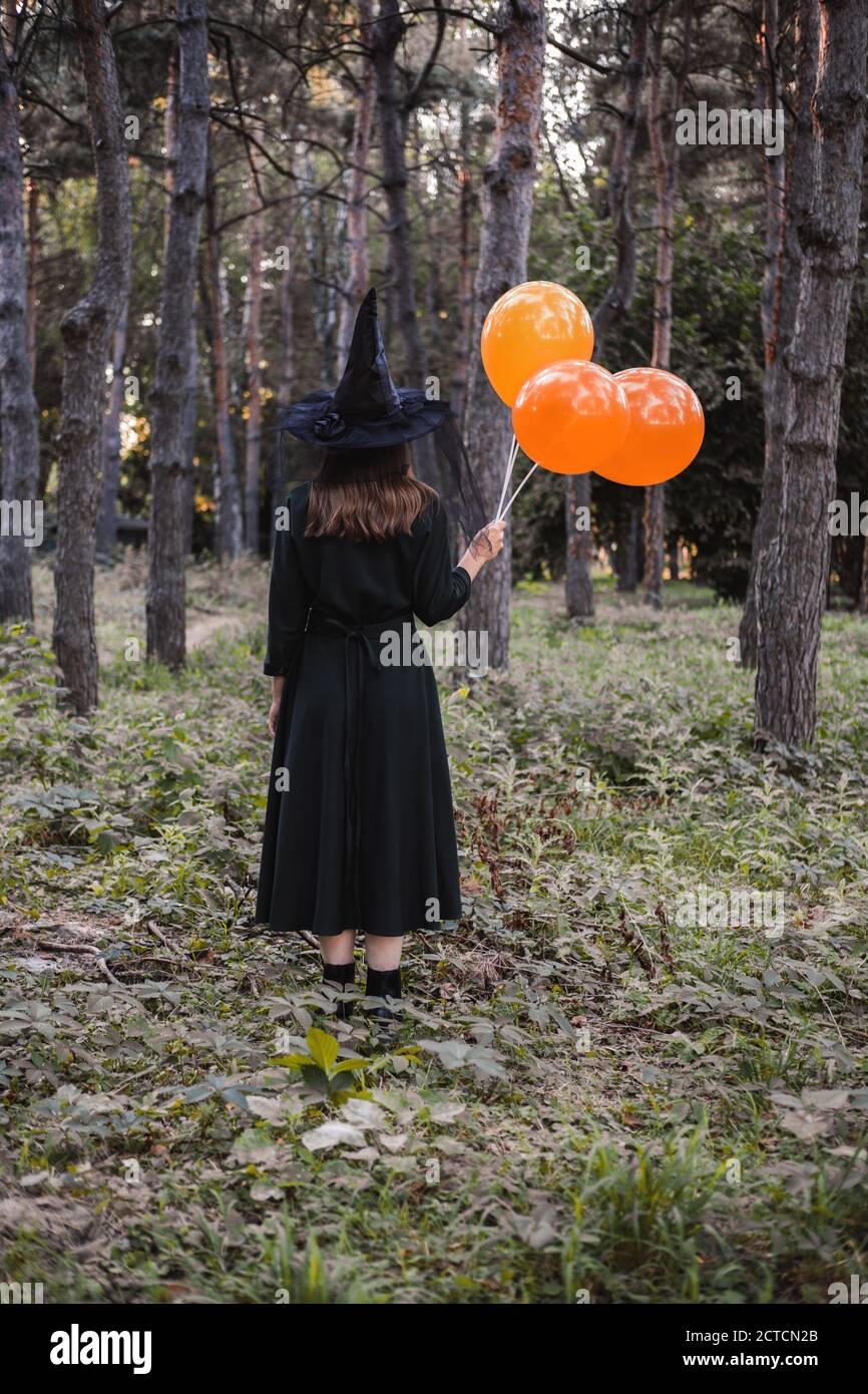 Jeune mignonne belle femme en robe sombre et chapeau de sorcière tient des ballons d'orange dans ses mains. Costume de fête d'Halloween. Forêt, parc avec arbres d'automne Banque D'Images