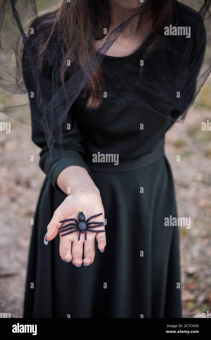 Jeune mignonne belle femme en robe sombre et chapeau de sorcière hotsblack araignée dans ses mains. Costume de fête d'Halloween. Forêt, parc avec arbres d'automne Banque D'Images