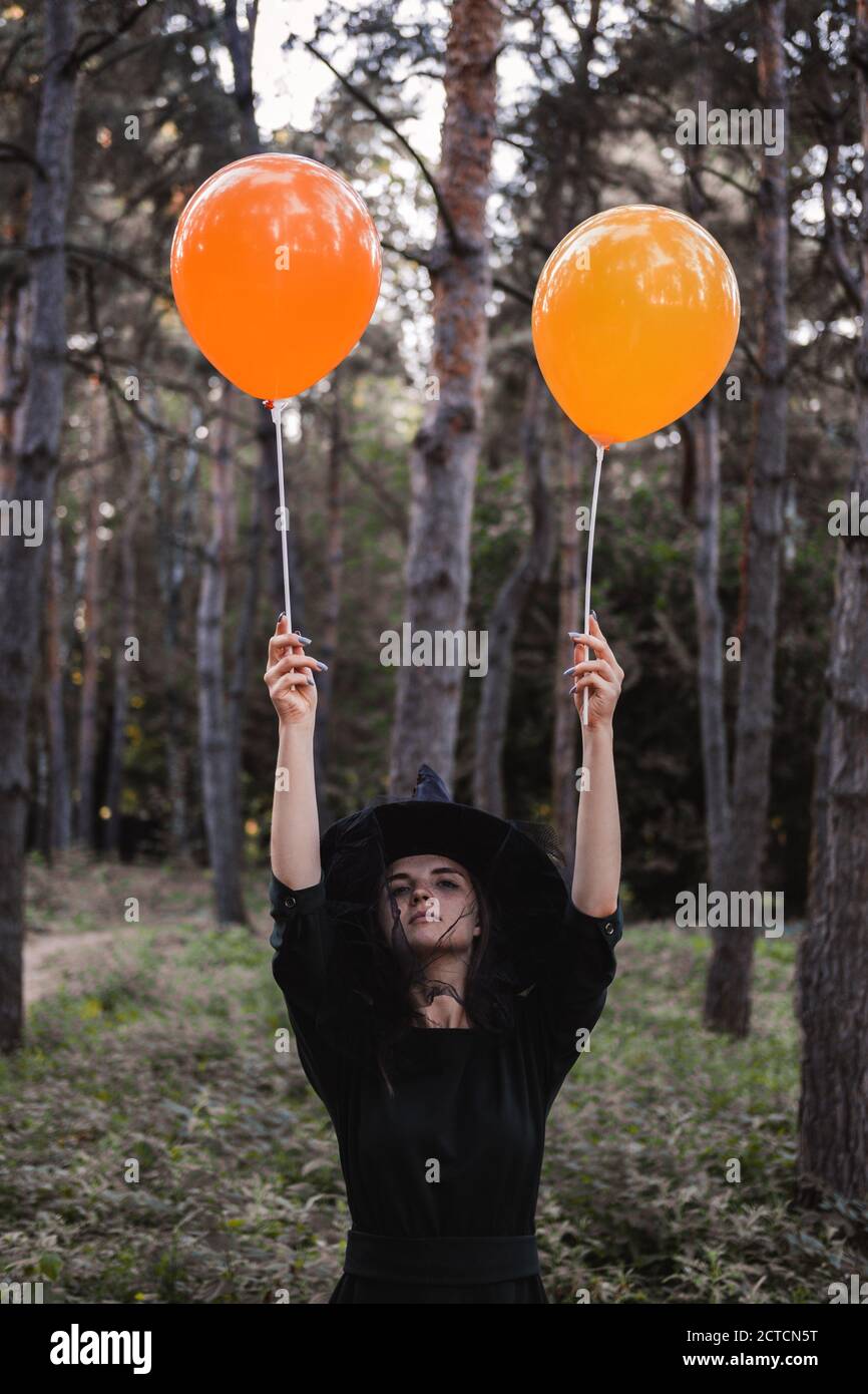 Jeune mignonne belle femme en robe sombre et chapeau de sorcière tient des ballons d'orange dans ses mains. Costume de fête d'Halloween. Forêt, parc avec arbres d'automne Banque D'Images