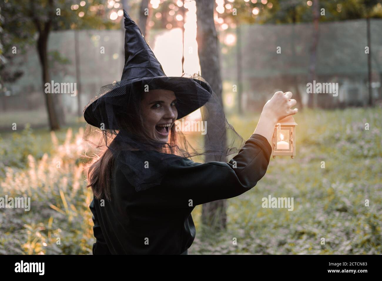 Une jeune femme en robe sombre et chapeau de sorcière tient une lanterne avec des bougies dans ses mains et illumine la forêt. Costume de fête d'Halloween. Garez-vous avec l'autom Banque D'Images