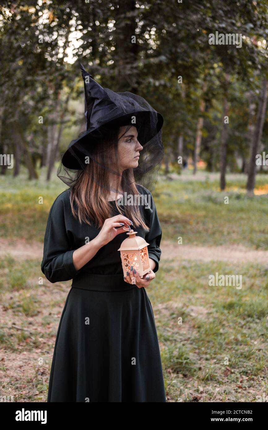 Une jeune femme en robe sombre et chapeau de sorcière tient une lanterne avec des bougies dans ses mains et illumine la forêt. Costume de fête d'Halloween. Garez-vous avec l'autom Banque D'Images