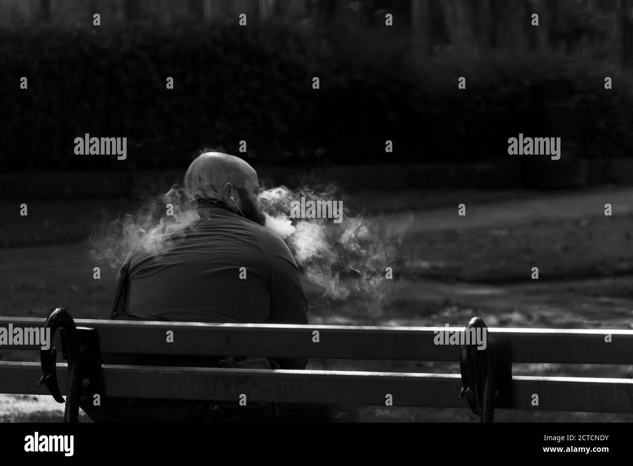Un gars s'est assis sur un banc de parc en bois de vapotage et de puffrage d'un nuage de fumée, Harrogate, North Yorkshire, Angleterre, Royaume-Uni. Banque D'Images