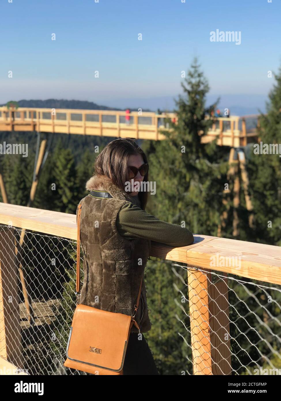 Jeune femme avec de longs cheveux bruns en fourrure gilet et avec sac à main orange dans des lunettes de soleil penchées sur une rambarde en bois d'un trottoir Banque D'Images