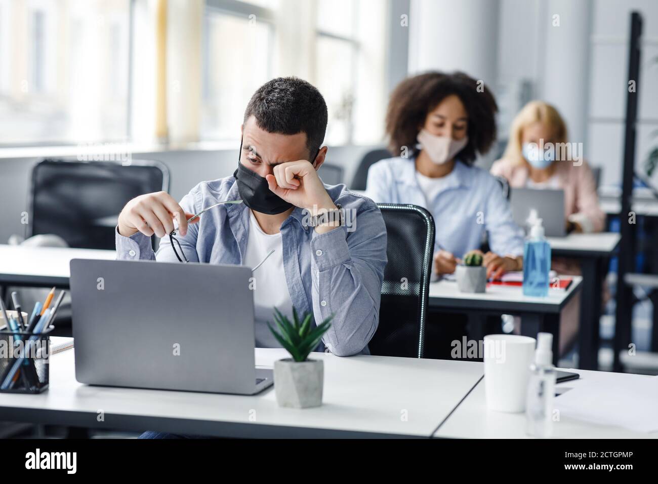 Fatigué et surtravaillé. Se concentrer sur l'homme dans le masque de protection frotte les yeux avec la main, derrière les collègues assis femmes sur les lieux de travail avec des ordinateurs portables et antiseptiques Banque D'Images