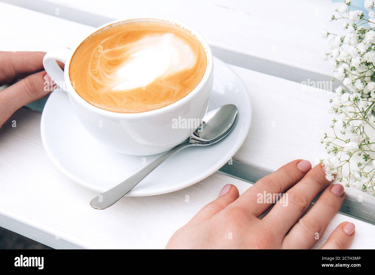 Mains de femmes et une tasse de cappuccino blanc en céramique, bouquet de petites fleurs blanches sur un banc en bois. Café rustique en plein air le matin. Routine du matin Banque D'Images