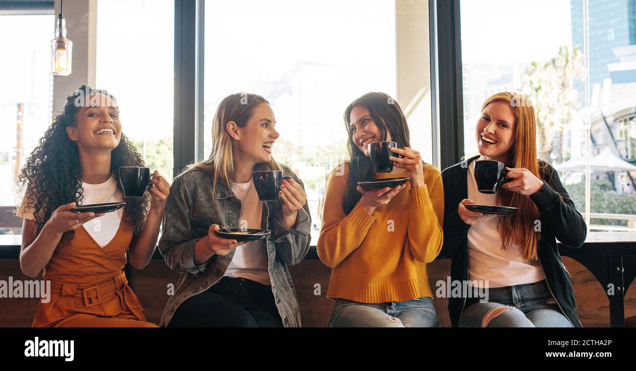 Des amies assises dans un café tenant une tasse de café et souriant. Groupe de quatre femmes prenant un café ensemble dans un café. Banque D'Images