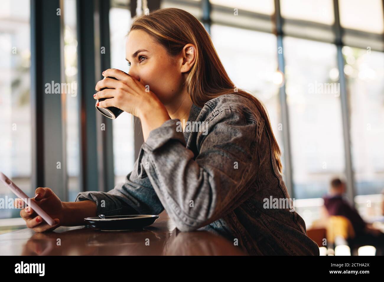 Femme dans un restaurant qui boit du café. Une femme assise dans un café avec un téléphone portable buvant une tasse de café. Banque D'Images