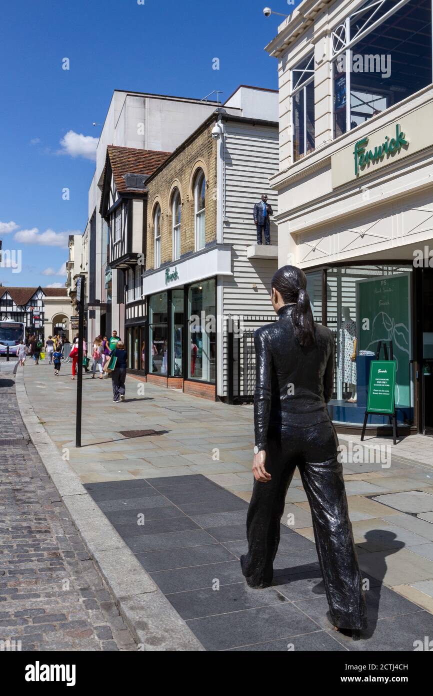 Une femme marchant le long de la rue, une des deux sculptures en bronze de Sean Henry, High Street, Colchester, Essex, Royaume-Uni. Banque D'Images