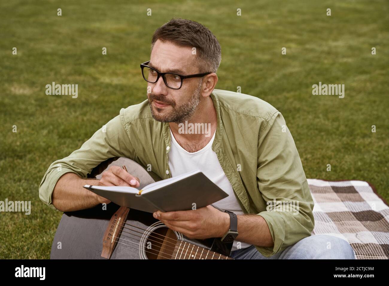 Jeune homme attentif portant des lunettes tenant une guitare acoustique et composant une chanson tout en étant assis sur une herbe verte à l'extérieur Banque D'Images