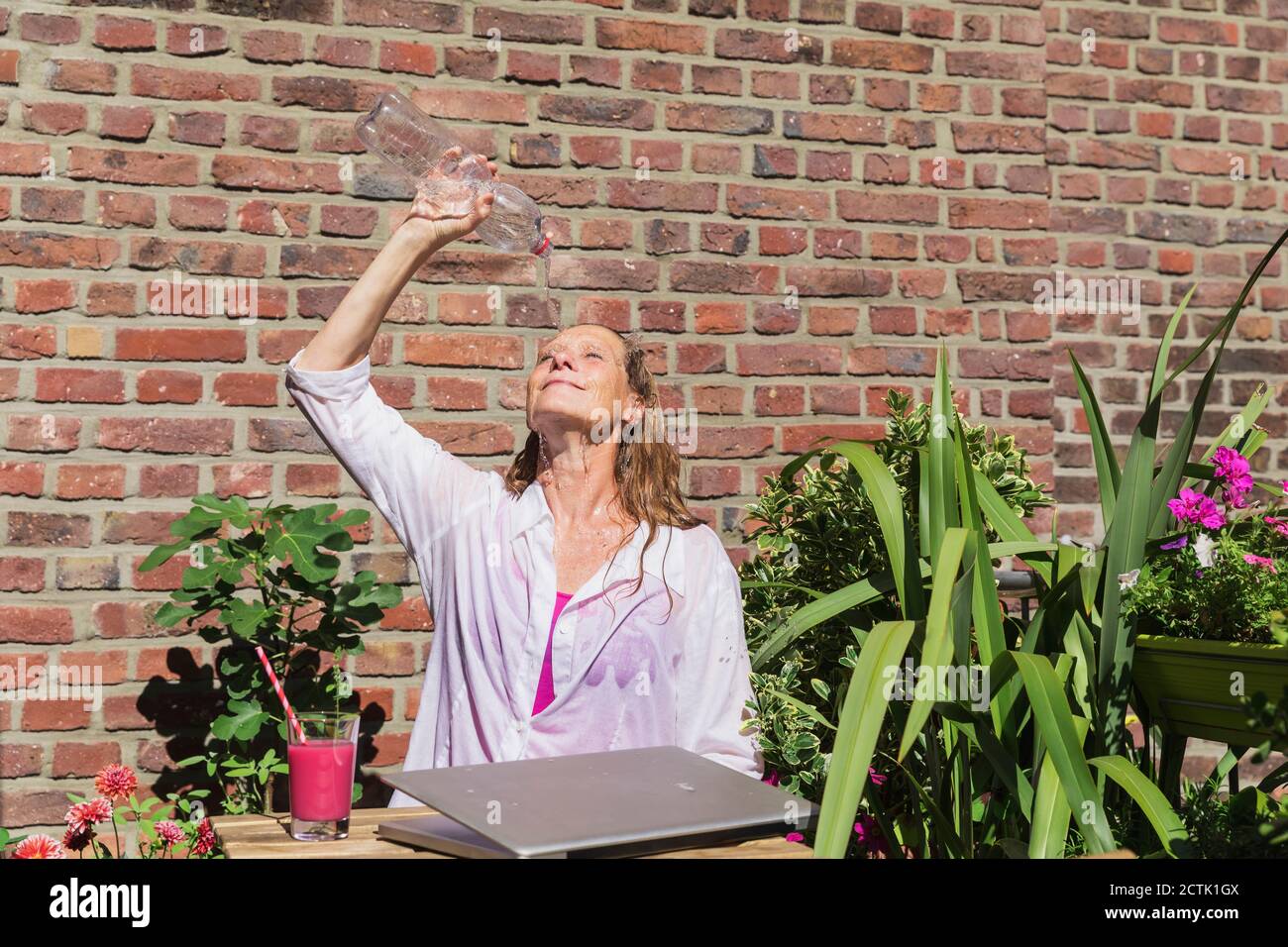 Femme d'affaires se rafraîchissant en versant l'eau de la bouteille sur le balcon contre le mur de briques pendant la pause le jour ensoleillé Banque D'Images