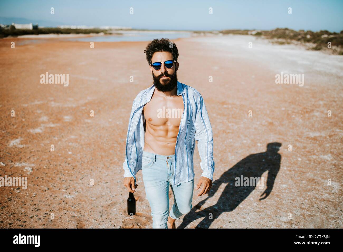 Jeune homme avec chemise ouverte marchant dans le désert tout en tenant bouteille de bière à la main le jour ensoleillé Banque D'Images