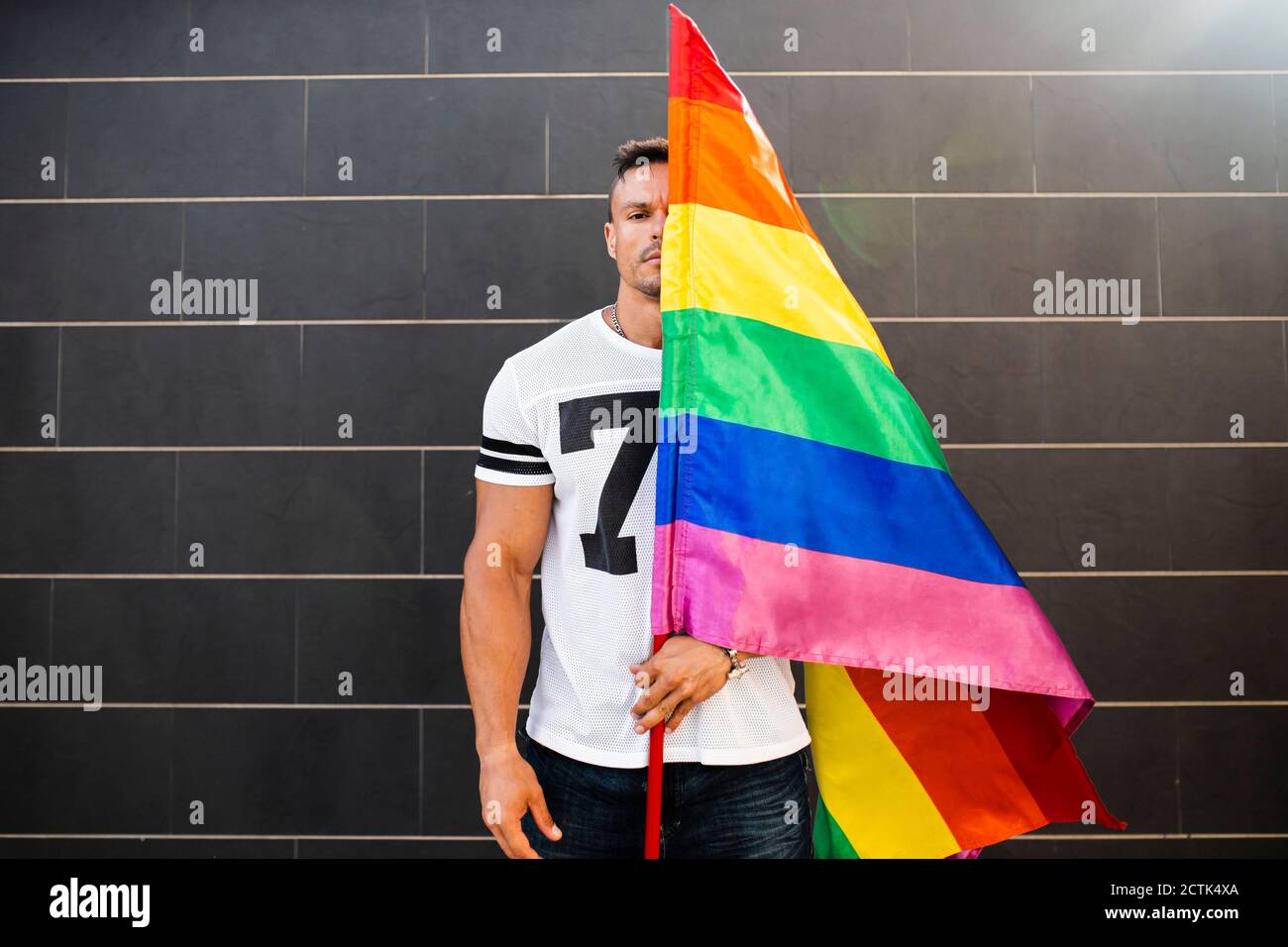Jeune homme tenant le drapeau arc-en-ciel tout en se tenant contre le mur Banque D'Images