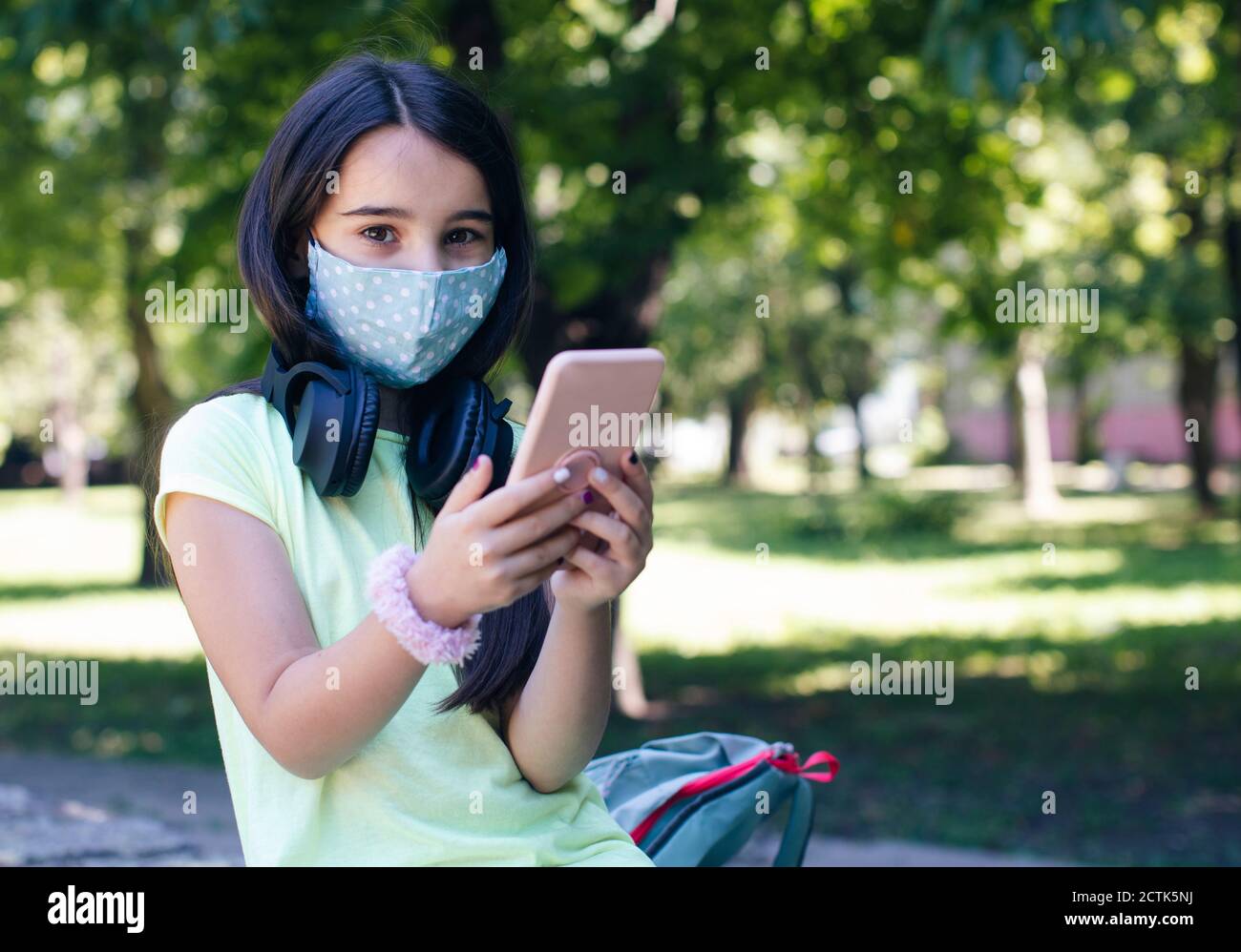 Fille avec masque facial utilisant un smartphone dans un parc public Banque D'Images