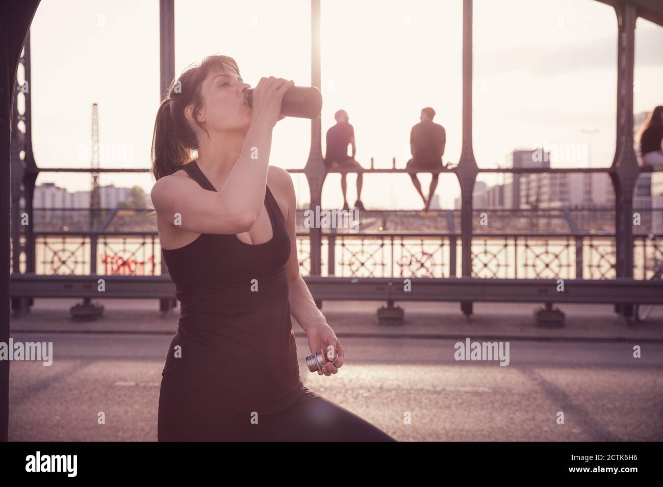 Femme buvant de l'eau de la bouteille après avoir fait de l'exercice sur le pont à coucher de soleil en ville Banque D'Images