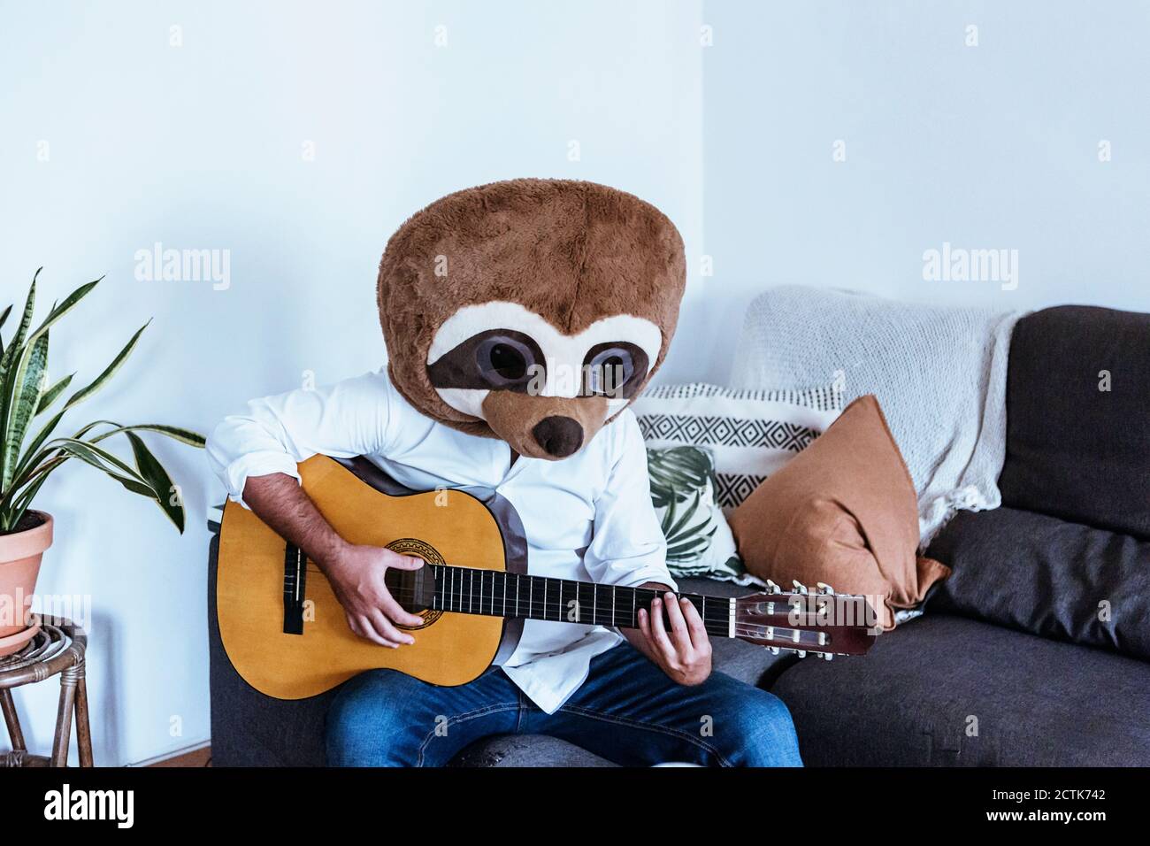 Homme adulte moyen portant un masque d'ours en peluche jouant de la guitare pendant salon avec canapé Banque D'Images