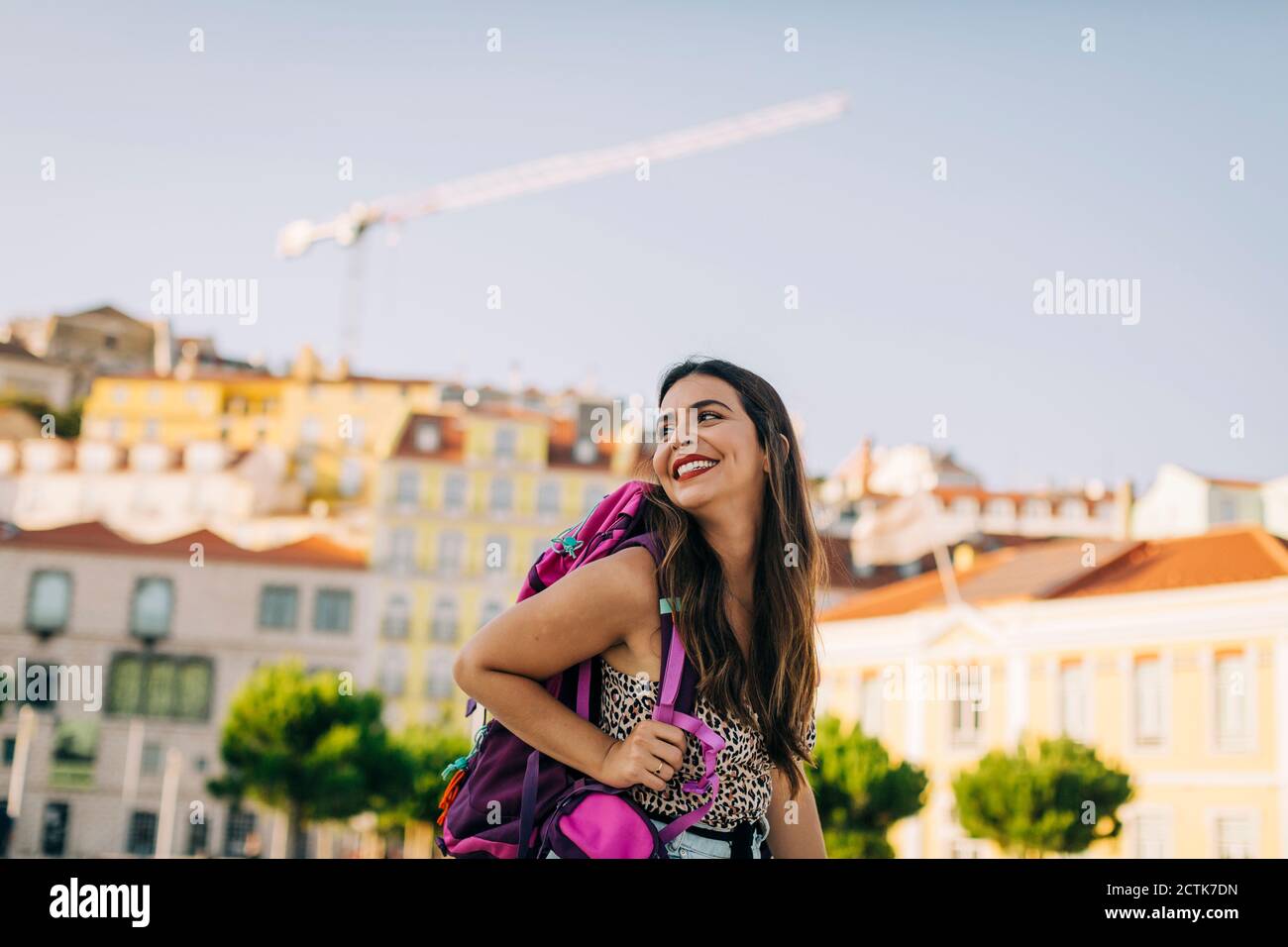 Jeune femme souriante avec sac à dos qui regarde loin tout en restant debout ville Banque D'Images