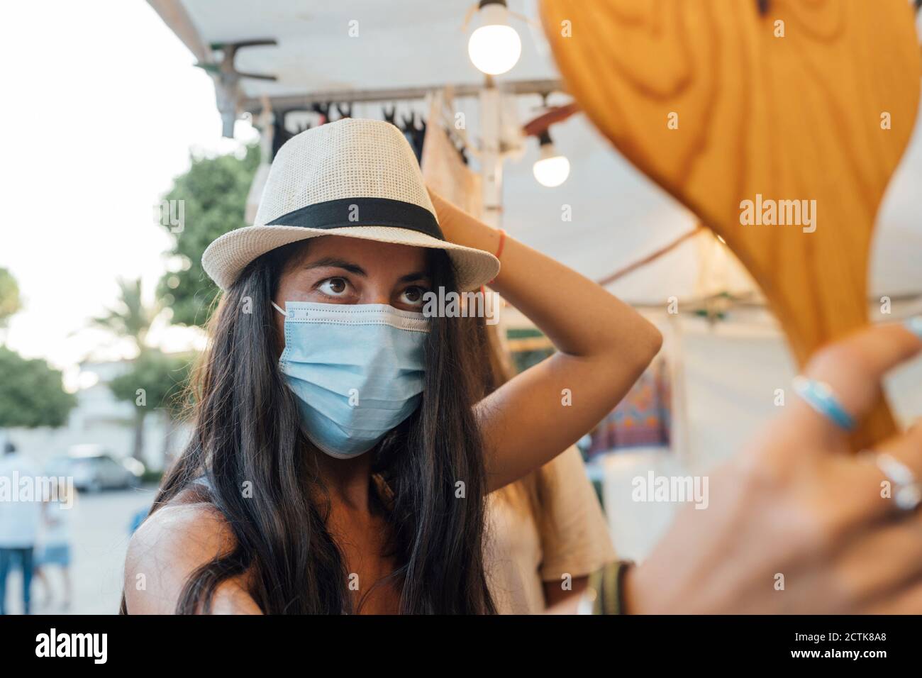 Femme portant un masque de protection pour le visage achetant un chapeau tout en regardant dans miroir sur la rue Banque D'Images