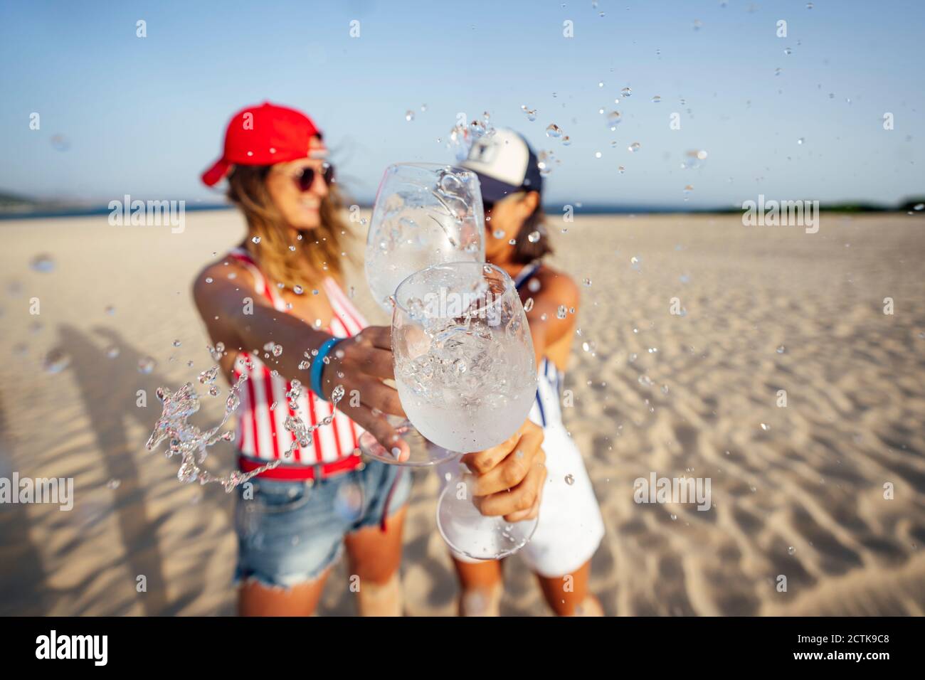 Des amis de sexe féminin de taille moyenne barbotent en buvant un verre tout en dorant à la plage par beau temps Banque D'Images