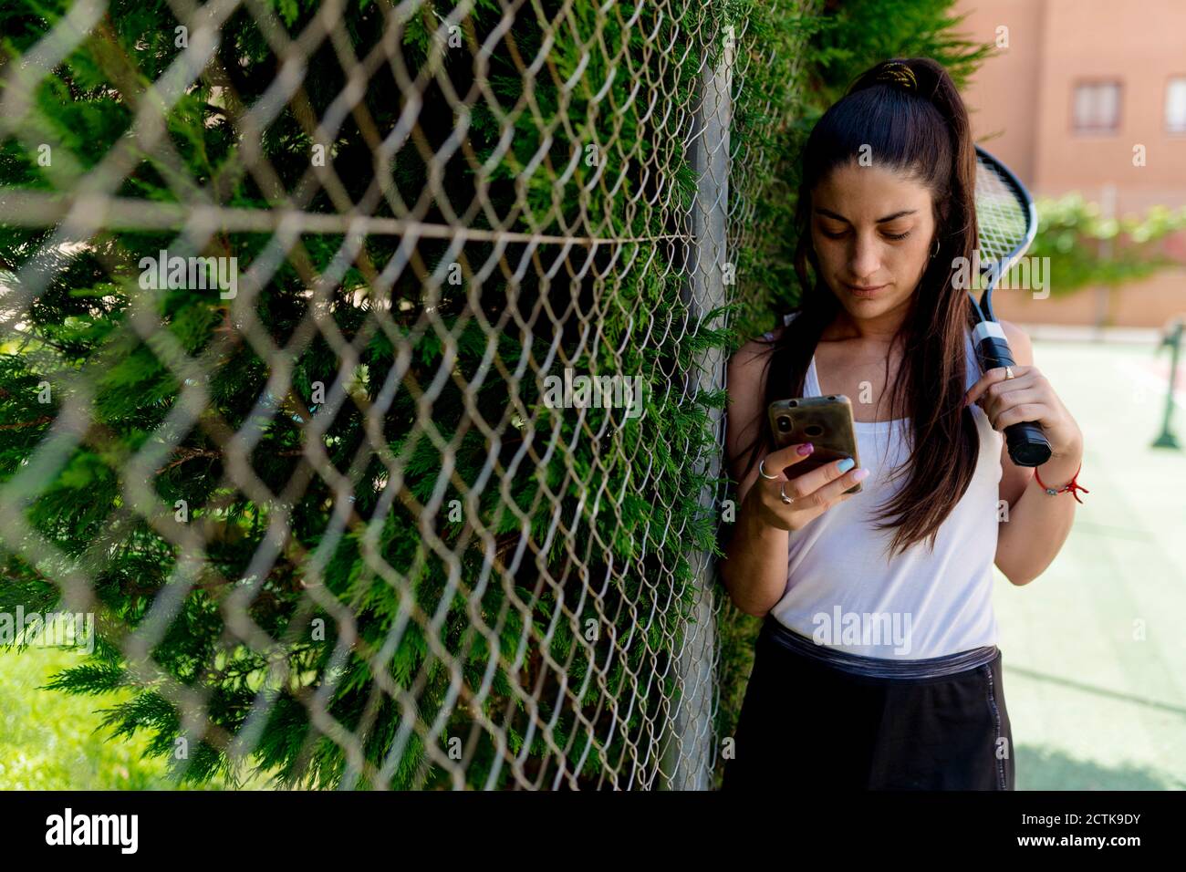 Joueuse de tennis féminine utilisant un smartphone en se tenant près d'une clôture en cour Banque D'Images